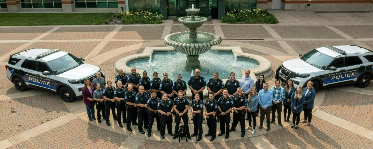 Group of police officers and some civilians posing in front of a statue for a group photo taken from above with two police cruisers on either side